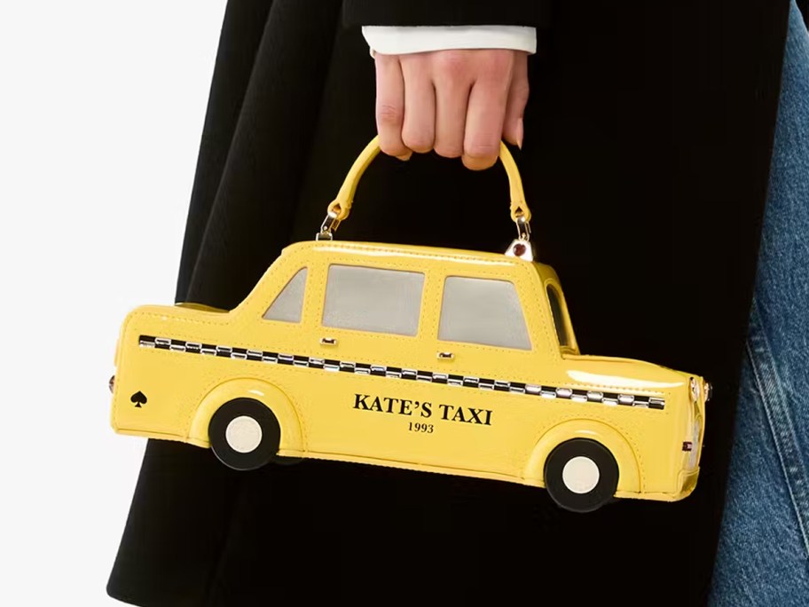 Person holding a Kate Spade New York 3D taxi crossbody bag shaped like a yellow cab with "KATE'S TAXI 1993" printed on the side.