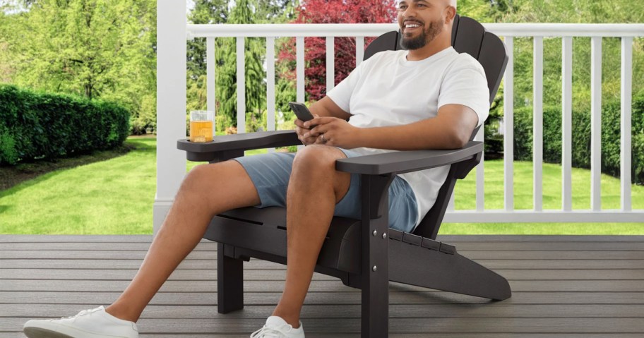 Man relaxing in a dark brown Keter Everest Adirondack Chair with built-in cupholder, seated on a porch with greenery in the background.