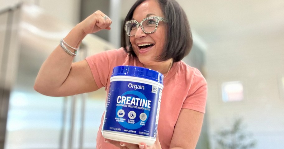 A smiling woman in glasses flexes her arm while proudly holding a large container of Orgain Creatine, standing in a bright, modern kitchen.