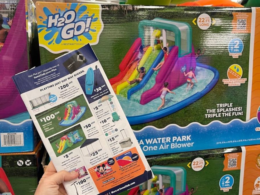 Hand holding a Sam's Club flyer advertising a sale on an H2O Go Water Slide in front of the box for the water slide