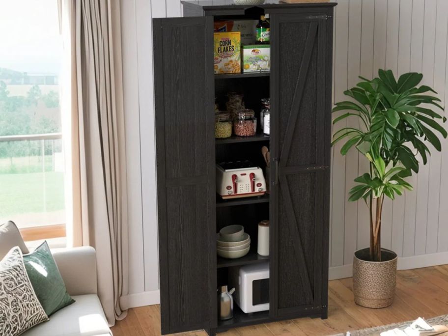 A black Gracie Oaks Ceasar Tall Storage Pantry Cabinet in a living room with one door open to show pantry items and small appliances inside