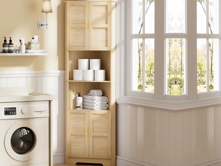 A Winston Porter Morden Tall Corner Cabinet in a bathroom with toiley paper and other bathroom essentials on the shelves