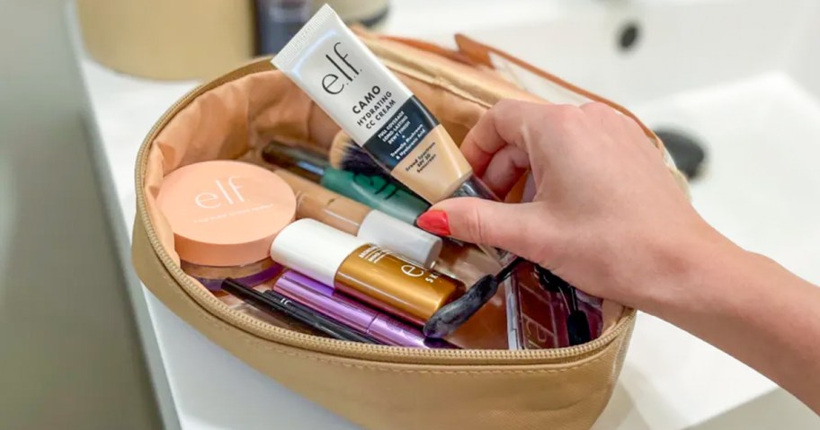 Open makeup bag filled with e.l.f. Cosmetics products, including Camo Hydrating CC Cream, setting powder, primer, and brushes, with a woman's hand reaching in.