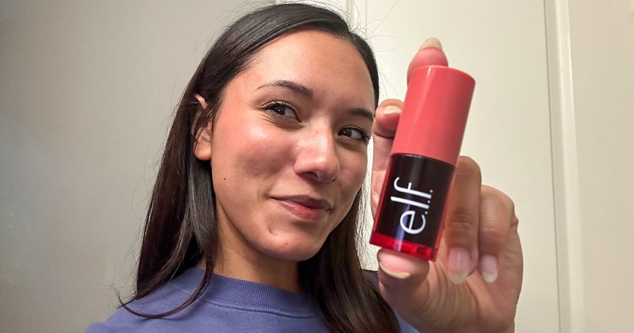 Woman holding e.l.f. Cosmetics blush tint with a coral cap, smiling and showing the product up close