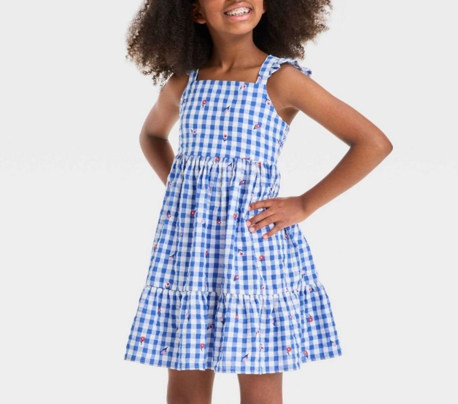 a little girl with her hands on her hips wearing a blue and white gingham dress with tiny floral print