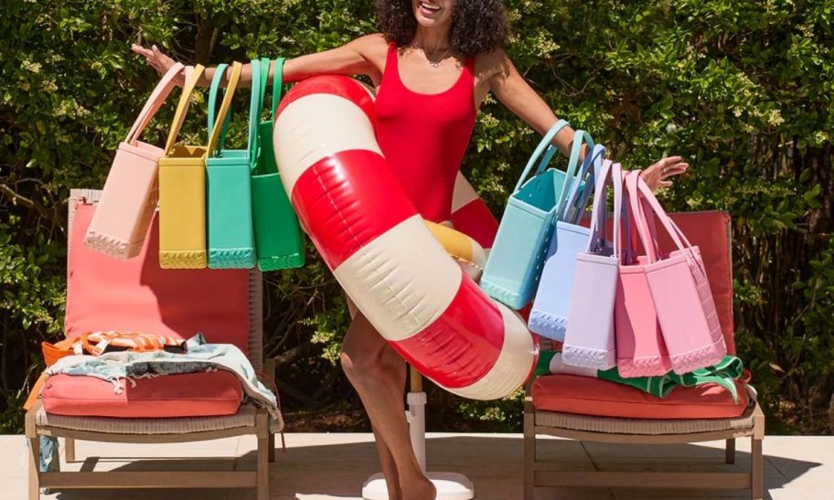 a woman in a red swimsuit and a red and white swim ring at poolside with arms out stretched and filled with bogg bags