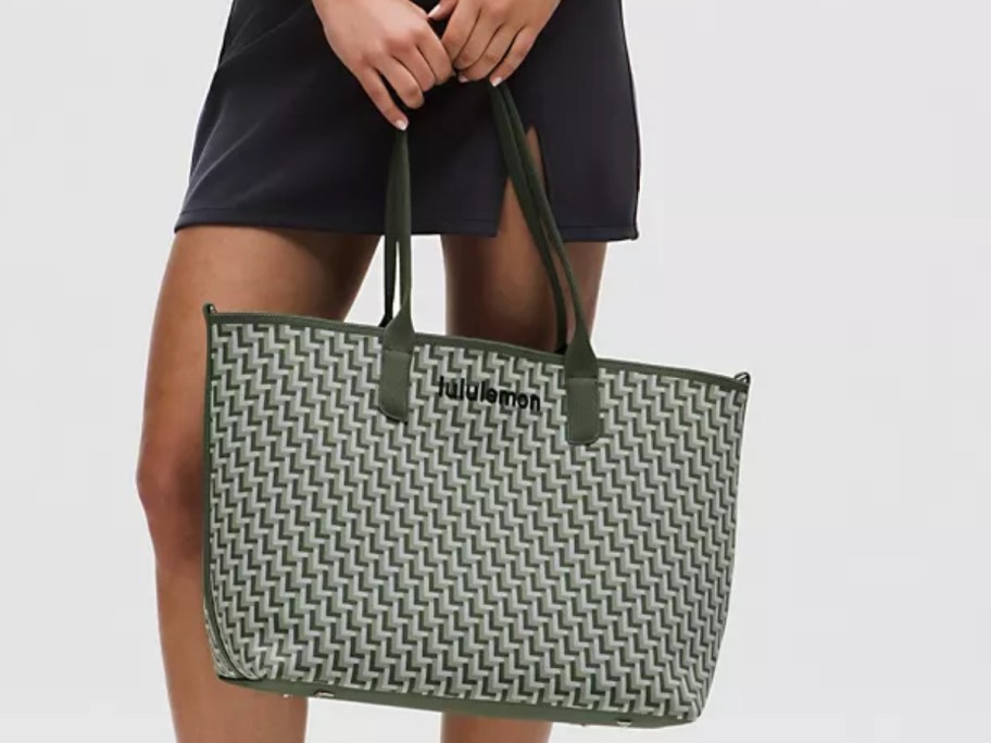 lower half of a woman wearing a black skirt and holding a large green and white knit tote bag