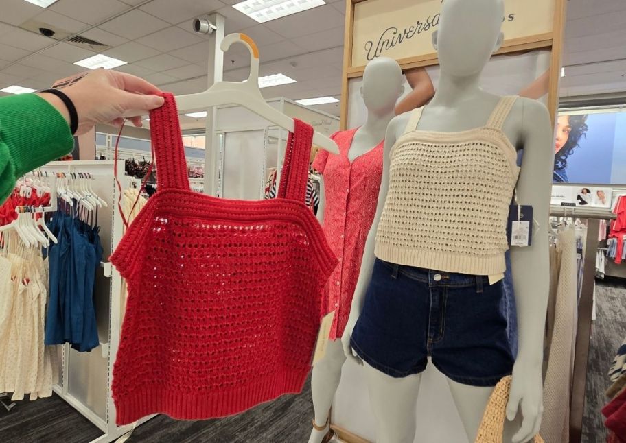 a womans hand holding a red crocheted tank on a hanger, next to an ivory crocheted tank on a mannequin