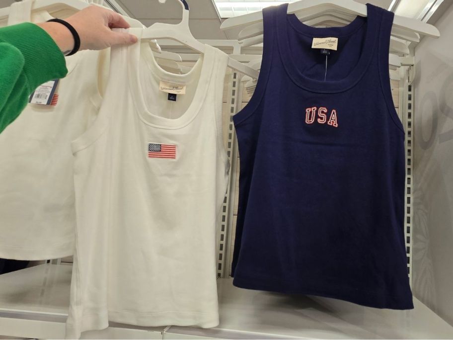a womans hand holding up a white american flag tank top next to a navy blue USA tank top hanging on a display wall. 