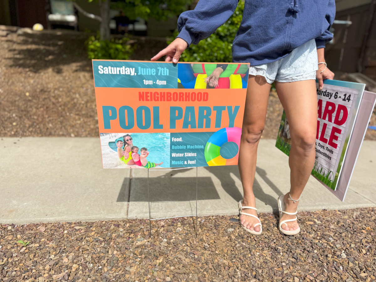 Woman standing next to custom pool party sign holding a yard sale sign in the other hand