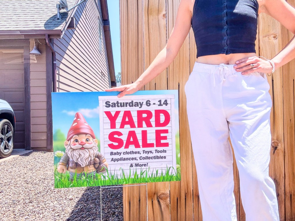 Woman wearing white pants and standing next to custom yard sale sign in front of fence