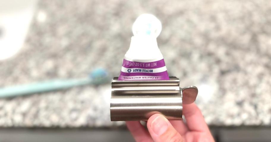A close-up of a toothpaste tube inserted into a metal tube squeezer, held by hand over a granite bathroom counter with a toothbrush in the background.