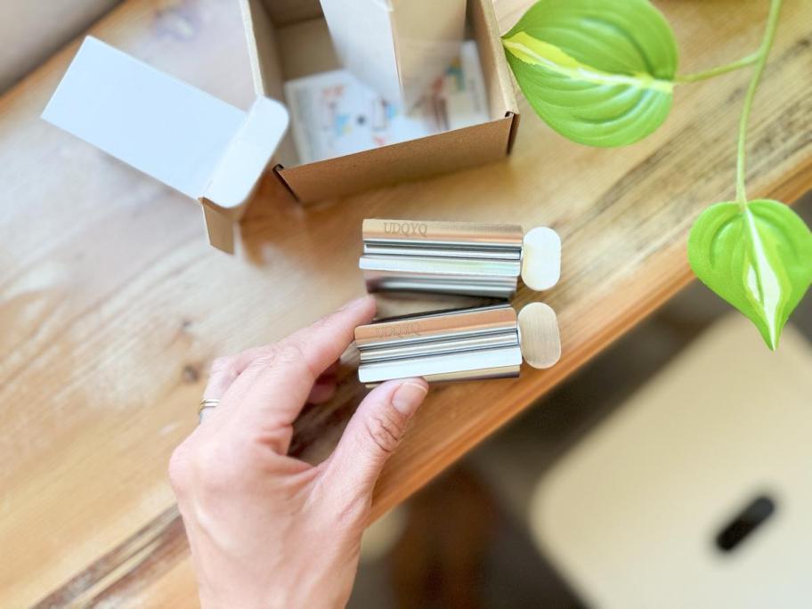A hand holding two metal tube squeezers on a wooden desk next to an open cardboard box and a green plant.
